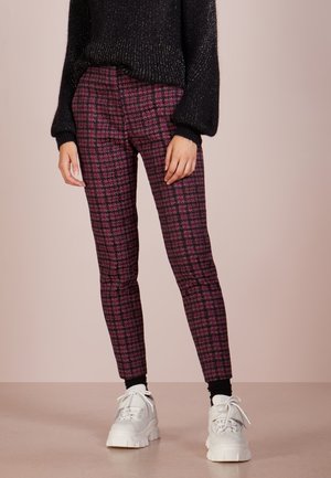 Persoon draagt nauwsluitende paarse en zwarte geruite broek, zwarte trui, zwarte sokken en witte chunky sneakers, staand op een beige vloer.