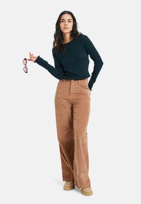 Navyblauwe blouse met lange mouwen en scalloped manchetjes gecombineerd met wijde bruine Corduroy-broek en beige plateauschoenen. Houdt een paar roze zonnebrillen vast.