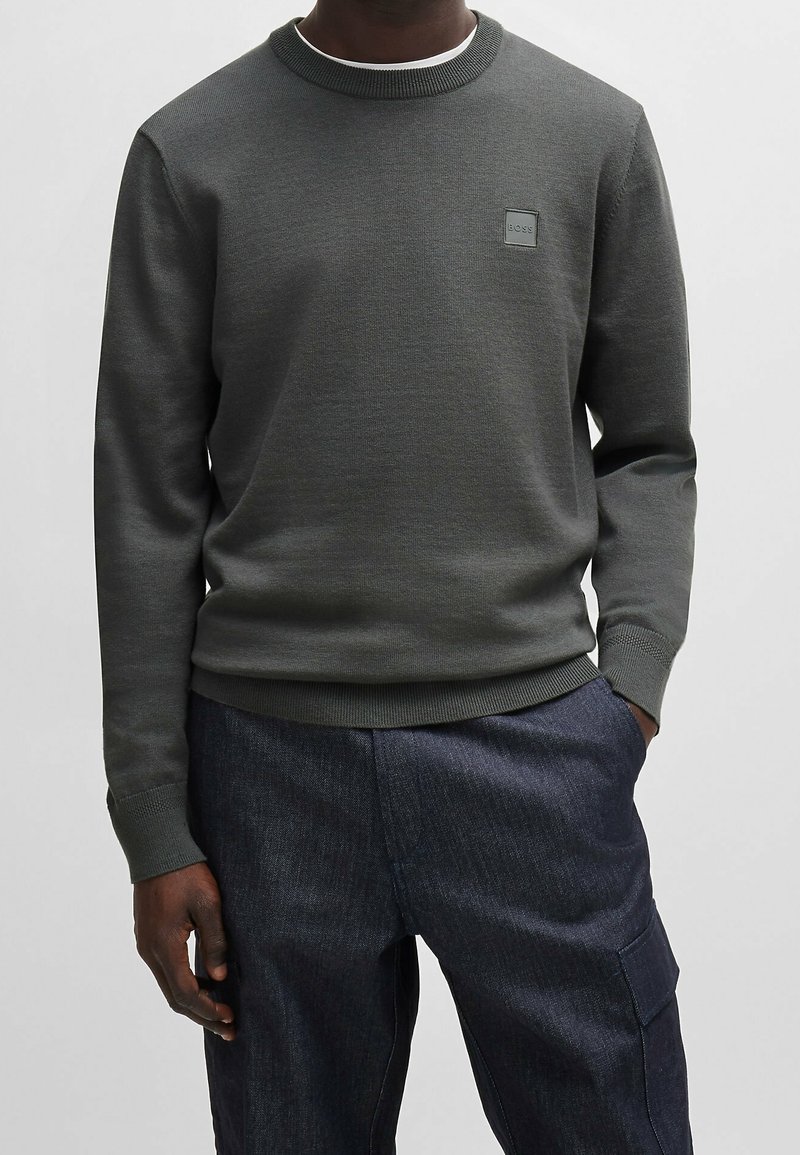 Homme portant un pull à manches longues gris foncé avec le logo BOSS sur la poitrine et un pantalon cargo bleu marine, debout avec une main dans la poche.