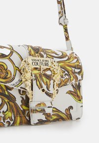 Vit handväska med guldfärgade detaljer som har intrikata bruna och gula barockmönster. Den har en axelrem och ett klaffstängningssystem.