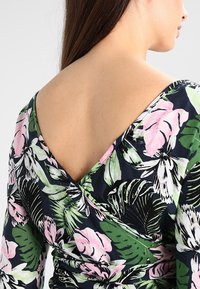 Haut à motif floral avec un fond bleu marine et des designs tropicaux roses et verts, décolleté en V profond et manches longues. Texture de tissu lisse.