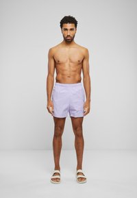 Pantaloni scurți de baie pentru bărbați, în nuanță de violet deschis, cu talie elastică, buzunare laterale și o croială lejeră, asortați cu sandale bej tip slide.