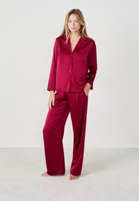 Burgundowy satynowy zestaw piżamowy z długim rękawem, zapinana na guziki bluzka z kieszonką oraz pasujące spodnie o luźnym kroju. Gładka tekstura.