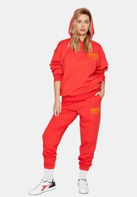 Completo di felpa rossa con cappuccio e pantaloni sportivi realizzati in tessuto morbido. Presenta un cappuccio, maniche elastiche e dettagli con logo stampato in giallo. Indossato con delle sneaker.