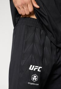 Μαύρο αθλητικό σορτς με υφή σχεδίου και ελαστική μέση. Λογότυπα UFC και crypto.com τυπωμένα με λευκό χρώμα στο μπροστινό μέρος.