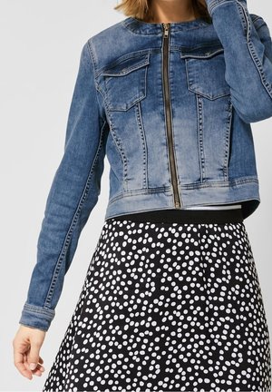 Frau trägt eine blaue Jeansjacke mit Reißverschluss vorne über einem schwarzen Rock mit weißem, unregelmäßigem Tupfenmuster.