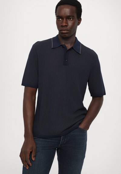 Marineblå kortermet polo-skjorte med ribbet tekstur, kontrasterende krage og treknappsstolpe. Brukt med mørke jeans.