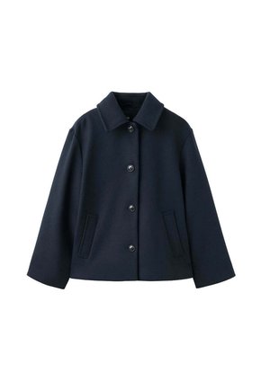Jeune femme portant une veste noire, un pull bleu, une chemise blanche, un jean large bleu clair et des chaussures noires, debout avec une main dans la poche.
