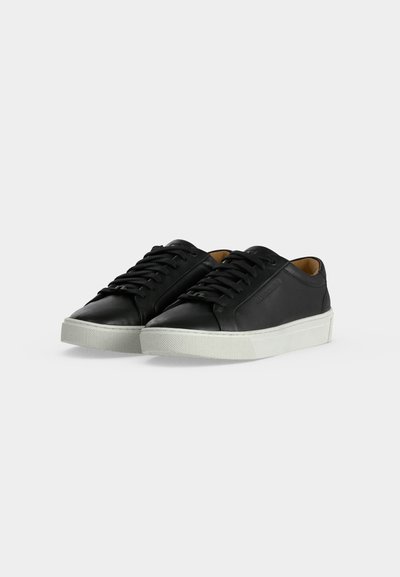 Ζευγάρι μαύρα δερμάτινα χαμηλά sneakers με λευκές λαστιχένιες σόλες και μαύρα κορδόνια σε απλό ανοιχτό φόντο.