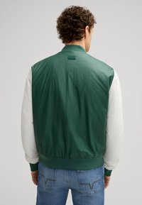 Giacca bomber color-block verde e bianca con polsini e fondo elasticizzati; texture liscia; presenta un logo sul retro. Indossata con jeans blu.