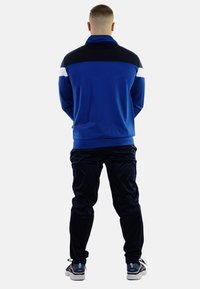 Blauwe zip-up jas met zwart-witte accenten, gecombineerd met zwarte broek en blauwe sneakers, gezien vanaf de achterkant. Soepele stofstructuur.