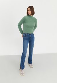 Maglione lungo a maniche lunghe di un verde chiaro e soffice, abbinato a jeans blu con spacchi laterali all'orlo, completato da sneakers chiare.