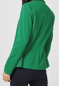 Zielona marynarka o dopasowanym kroju, z wyraźnymi ramionami i szwami na plecach podkreślającymi sylwetkę. Materiał wydaje się gładki i lekko elastyczny.