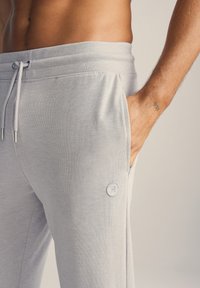 Lys grå joggerbukser lavet af blødt stof, med elastisk talje, sidelommer og et dekorativt logo-patch på låret.