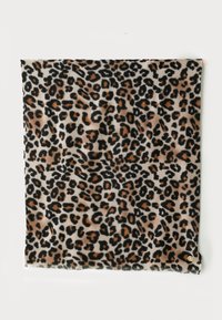 Carré de tissu à motif léopard avec des taches noires, brunes et beiges, suspendu contre un mur blanc, comportant une petite étiquette circulaire dorée.