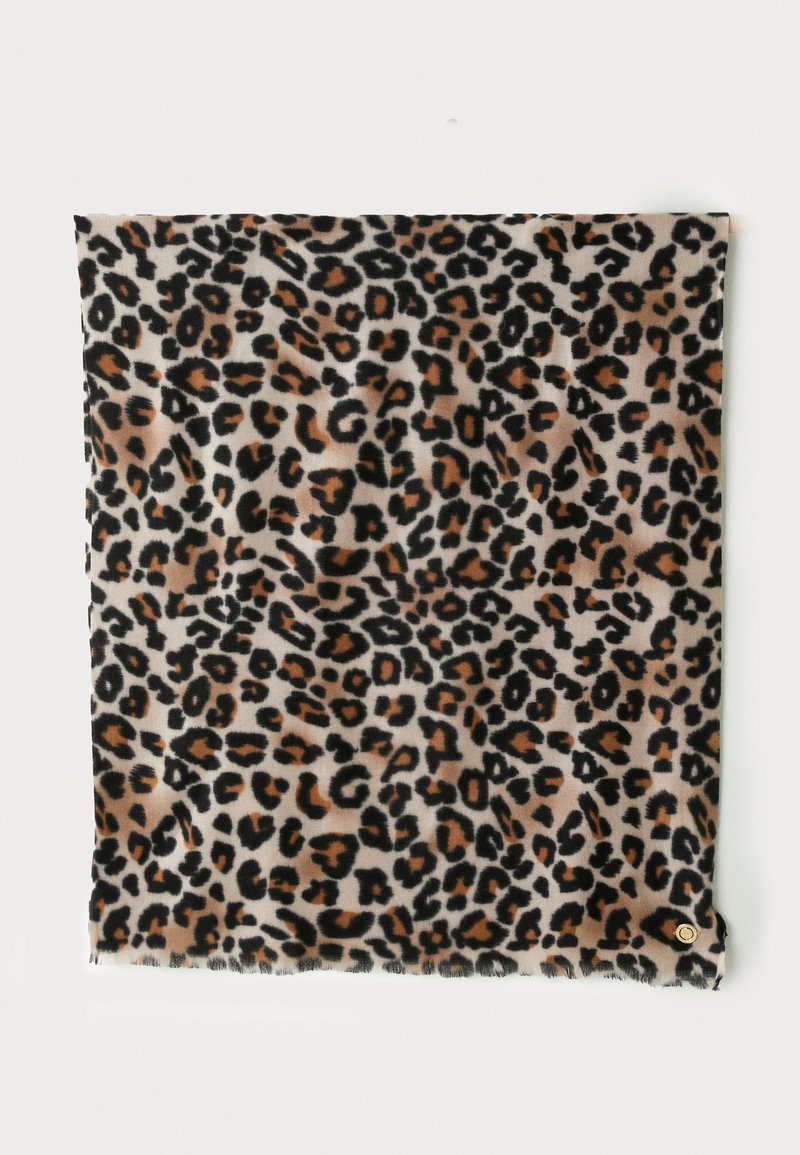 Carré de tissu à motif léopard avec des taches noires, brunes et beiges, suspendu contre un mur blanc, comportant une petite étiquette circulaire dorée.