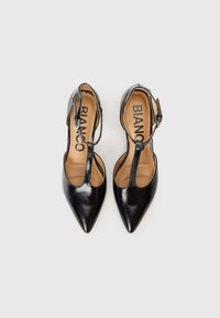 Scarpe alte in pelle nera con punte appuntite, design a T-strap e chiusura con fibbia; soletta color nocciola con lettering del marchio visibile.