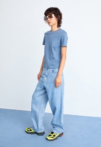 Blaues Baumwoll-T-Shirt mit kurzen Ärmeln, lockere hellblaue Jeanshose und schwarze sowie gelbe Sneaker. Das Modell trägt übergroße Sonnenbrillen.