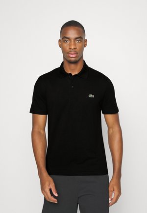 Polo noir en coton, avec une patte de boutonnage, des manches courtes et un petit logo crocodile vert sur la poitrine gauche.