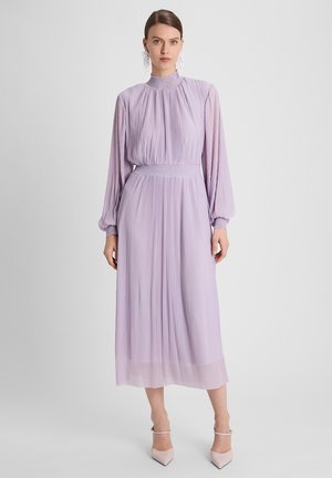 Vrouw draagt een lavendelkleurige midi-jurk met plooien, lange mouwen en een hoge hals, met een gerimpelde taille en manchetten, gecombineerd met lichtroze puntige hakken.