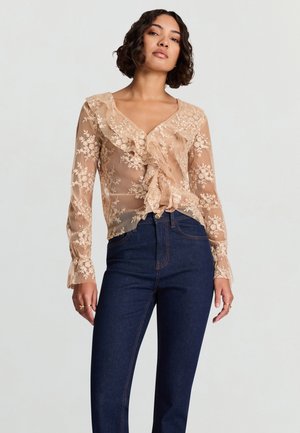 Camicia beige trasparente con dettagli in pizzo floreale, scollo arricciato, maniche lunghe e fiocco frontale. Abbinata a jeans a vita alta di colore blu scuro.