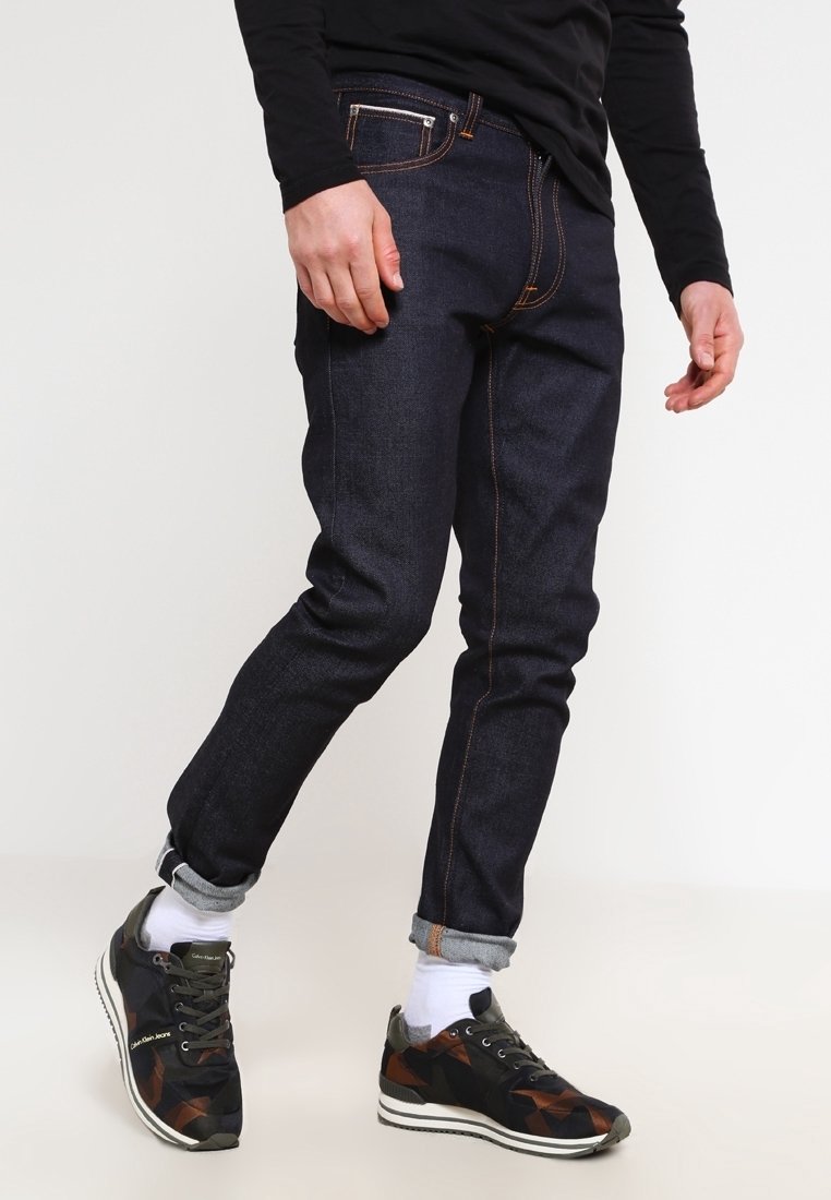 Mörkblå denimjeans med orange sömmar, slim fit-design och uppvikta fållar. Bärs med sneakers i kamouflage-mönster och vita strumpor.