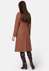 Brązowy wełniany płaszcz z paskiem w talii, długimi rękawami, rozporkiem z tyłu i bocznymi kieszeniami. Zestawiony z brązowymi skórzanymi botkami do kolan z marszczeniem.
