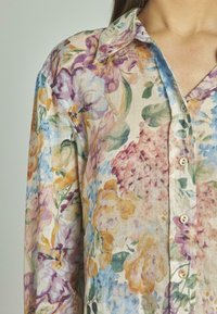 Camisa de botón floral en tejido ligero. Presenta patrones florales multicolores con matices de púrpura, azul, amarillo y verde.