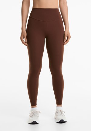 Höga bruna leggings gjorda av elastiskt tyg med en slät yta, kombinerat med ljusa sneakers och vita strumpor.