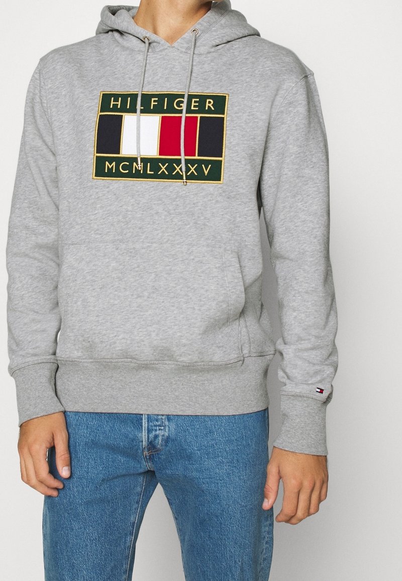 Hombre vestido con una sudadera gris con un logo rectangular multicolor y jeans azules, de pie contra un fondo claro y liso.