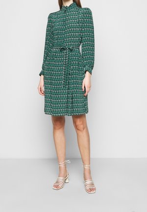 Vestido verde con estampado floral, cuello alto, mangas largas, se puede ajustarse en la cintura con un lazo. Combinado con sandalias de tiras color crema.