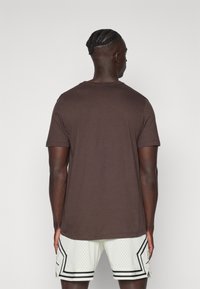 Man med flätor står vänd bort, iförd en enkel brun t-shirt med kort ärm och vita basketshorts med svarta detaljer.