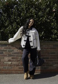 Hvid fleecejakke med sorte detaljer og knapper, båret over en mørk top, parret med mørke brede jeans og beige støvler; holder en sort håndtaske.