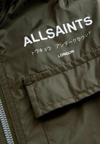 Olive zelená nylonová bunda s prednou kapsou, bielym potlačeným názvom značky "ALLSAINTS" a "LONDON," s textom v ďalších znakoch nižšie.