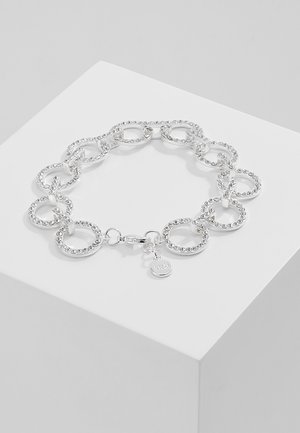 Bracelet en argent avec des chaînes circulaires liées ornées de strass et un petit charme rond sur un socle blanc.