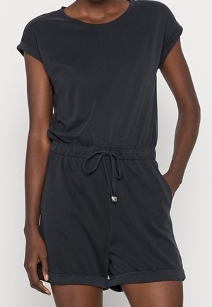 Nainen, joka käyttää mustaa, lyhythihaista haalaria, jossa on käännetyt kalvosimet, kiristysnauha vyötäröllä ja sivutaskut, seisoo yksinkertaisen taustan edessä.