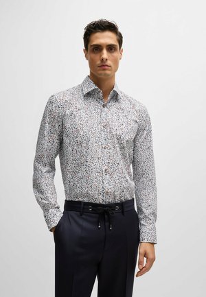 Chemise à manches longues, à boutons, avec un fond blanc, ornée d'un petit motif floral multicolore. Boutons marron et col évasé.