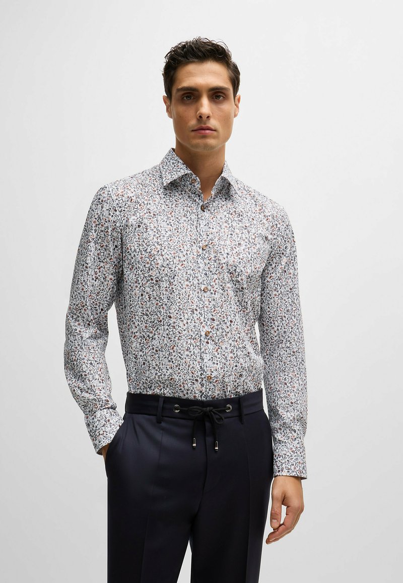 Chemise à manches longues, à boutons, avec un fond blanc, ornée d'un petit motif floral multicolore. Boutons marron et col évasé.