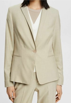 Blazer beige taillé avec un seul bouton, porté sur un top blanc et une chemise beige côtelée, assorti à un pantalon beige.