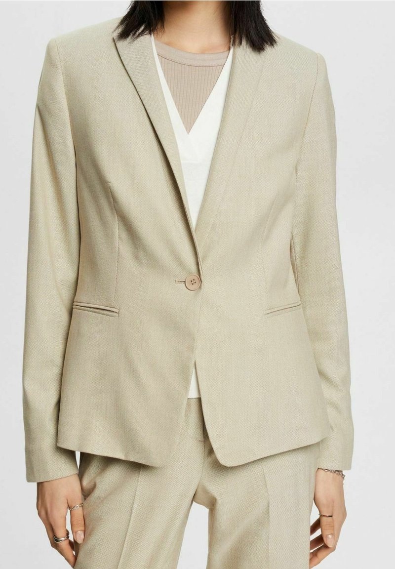 Blazer beige taillé avec un seul bouton, porté sur un top blanc et une chemise beige côtelée, assorti à un pantalon beige.