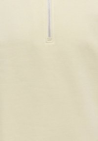 Gros plan sur un tissu blanc cassé avec une fermeture éclair argentée centrée, partiellement ouverte, montrant une couture soignée autour de la base de la fermeture éclair.