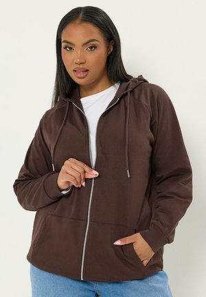 Frau mit brauner Kapuzenjacke mit Reißverschluss über einem weißen Shirt, mit einer Hand am Reißverschluss und der anderen in der Tasche, posiert vor einem einfarbigen Hintergrund.