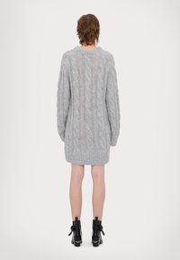 Personne debout, de dos, portant une robe-pull en maille torsadée gris clair et des bottines noires, sur fond blanc.
