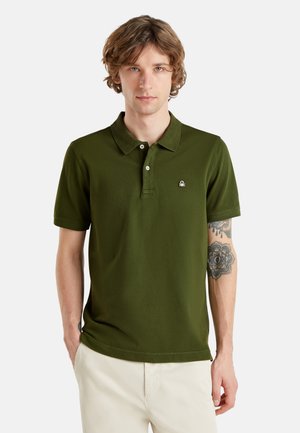 Młody mężczyzna z falowanymi włosami, ubrany w oliwkową, krótkim rękawem koszulkę polo i białe spodnie, pokazujący kwiatowy tatuaż na lewym przedramieniu.