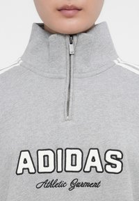 Close-up de uma pessoa a usar um sweatshirt cinzento da Adidas com um zíper de colarinho alto e o texto "Athletic Garment" abaixo do logotipo.