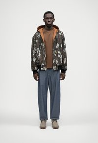 Braune gemusterte Bomberjacke mit einem beigen Innenfutter, kombiniert mit einem braunen T-Shirt und lockeren blauen Hosen. Beigefarbene Stiefel mit Gummisohle.