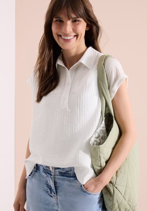 Lachende vrouw met een witte korte mouwen blouse en lichte denim jeans, die een lichtgroene doorgestikte schoudertas met een patroonvoering vasthoudt.