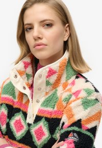 Maglione in pile colorato con motivo geometrico in rosa, verde e arancione, dotato di collo alto e bottoni a pressione. Logo del marchio sulla manica.