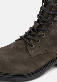 Cizma scurtă din suede maro, cu sireturi negre, orificii întărite, suprafață texturată și talpă din cauciuc negru. Prezintă cusături contrastante.