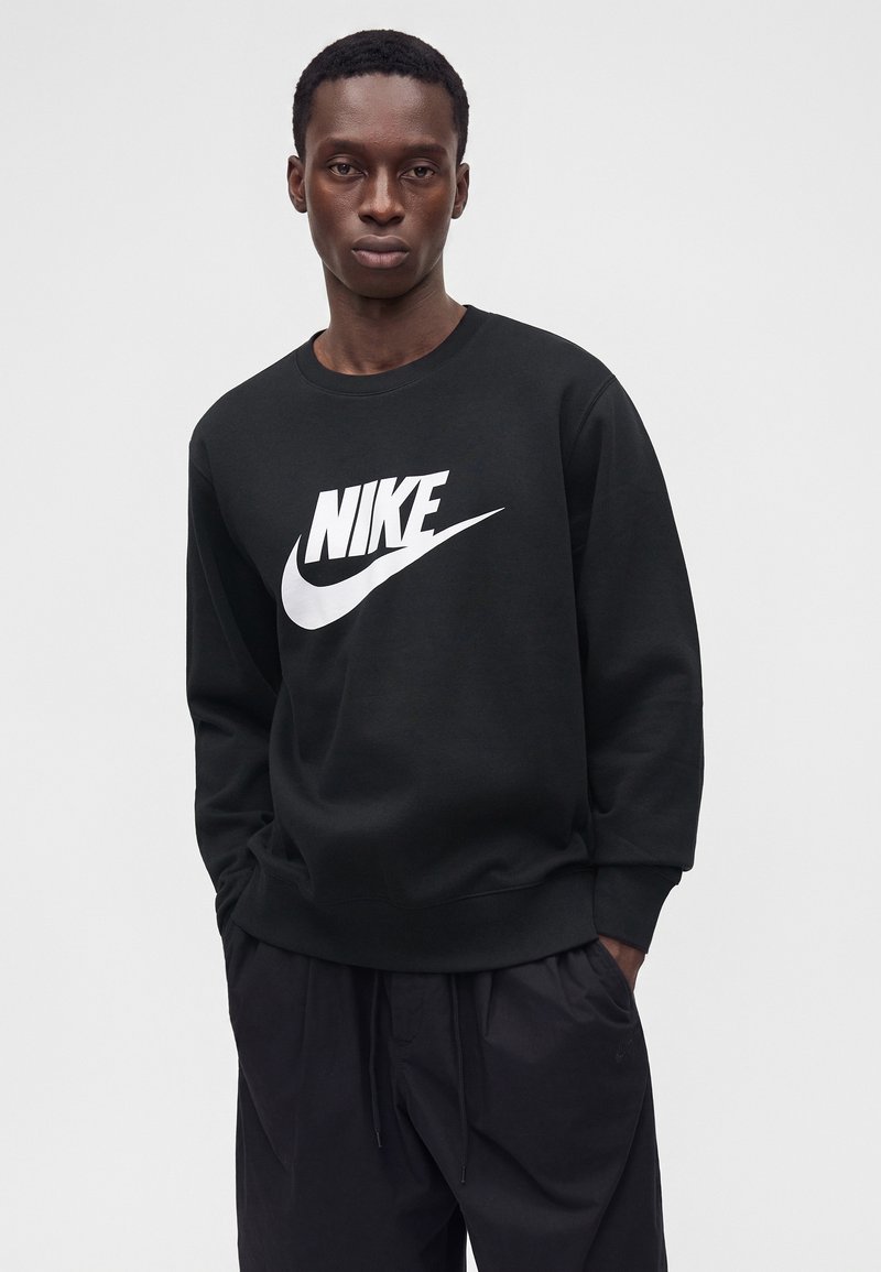 Joven con sudadera negra de Nike con logo blanco, de pie con las manos en los bolsillos frente a un fondo liso y claro.